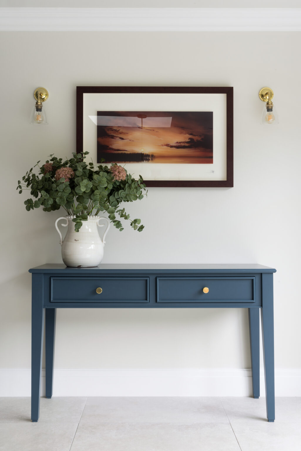 Deep blue console table with brass knobs, topped with a vase of foliage beneath a framed sunset print and wall lights
