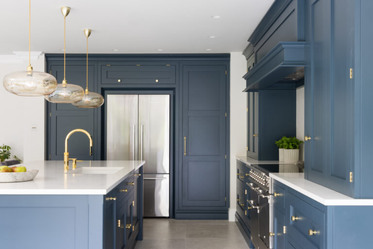 Deep blue kitchen cabinetry with brass handles, integrated fridge, range cooker and an island with brass tap and pendant lights
