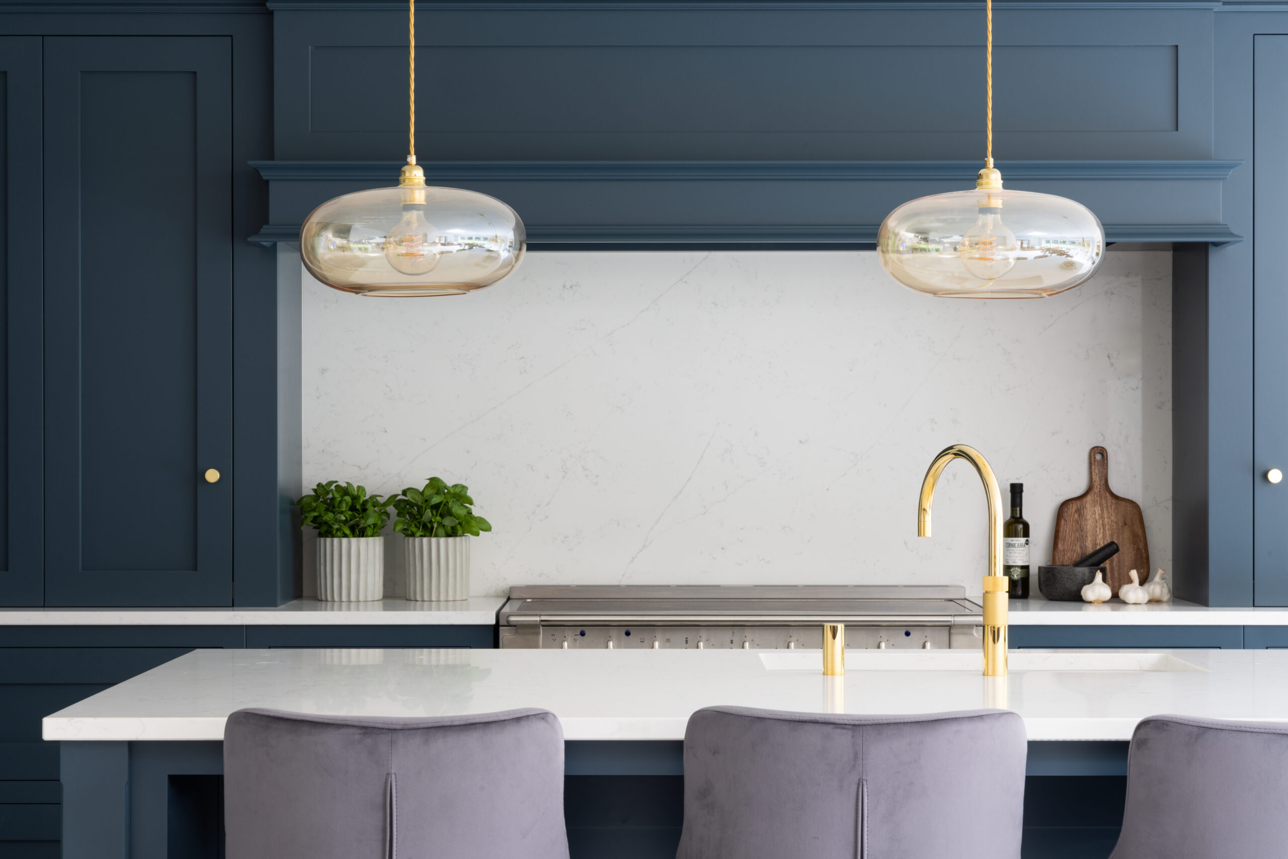 Deep blue kitchen cabinetry with brass pendant lights, a brass tap and velvet stools at the central island