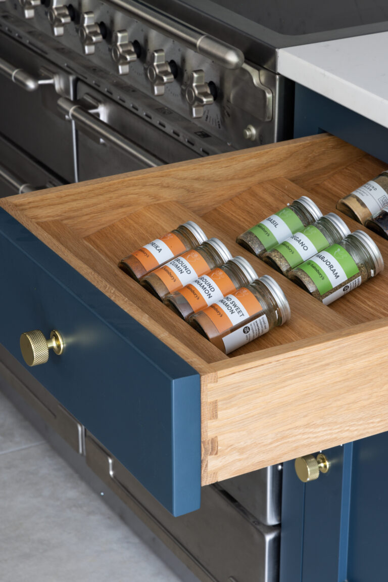 Open deep blue drawer with brass knob holding spice jars, beside a stainless steel range cooker