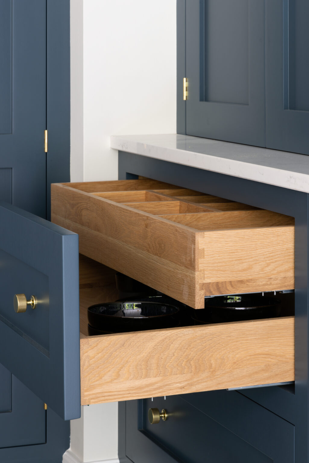 Open timber-lined drawer with sectioned insert beneath deep blue cabinetry and brass knurled handles