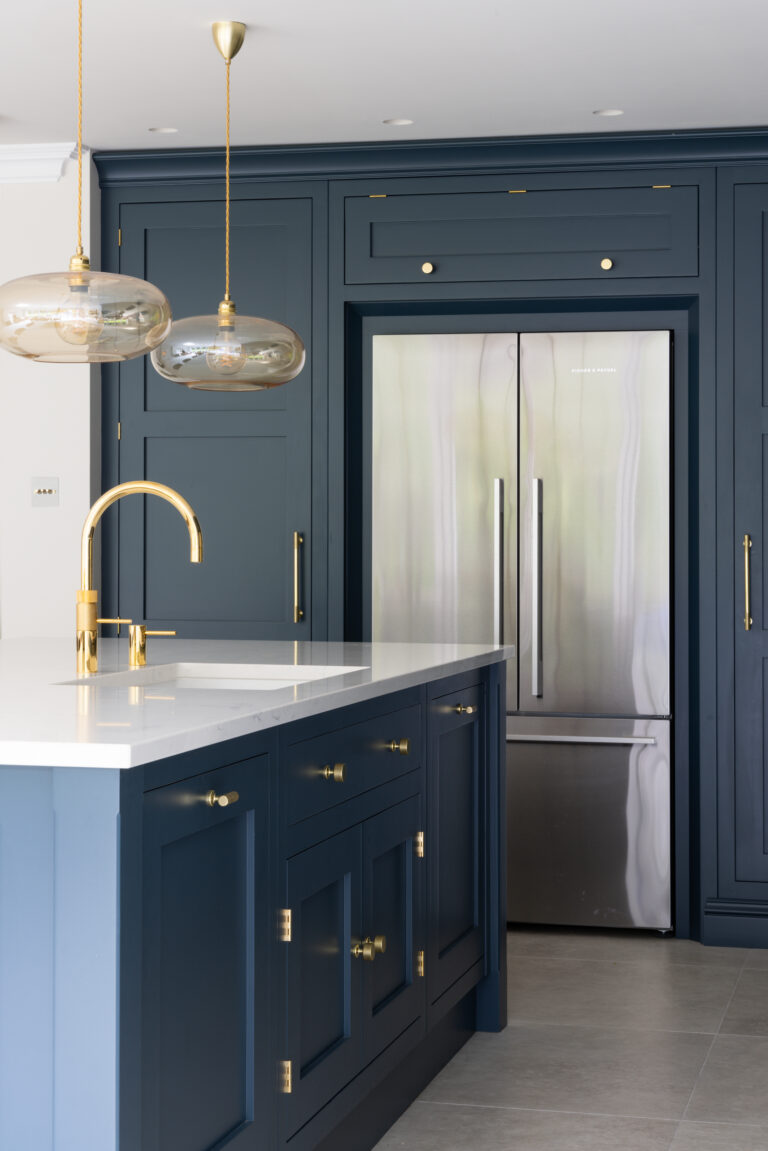 Deep blue kitchen cabinetry with brass handles and tap, an island with pale worktop, and an integrated American-style fridge freezer