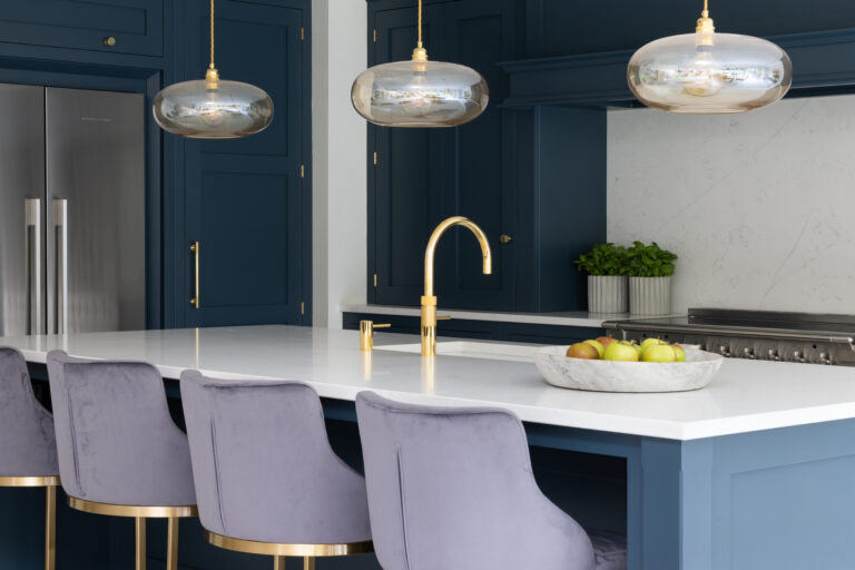 Kitchen island with velvet stools, brass tap and pendant lights, set against deep blue cabinetry
