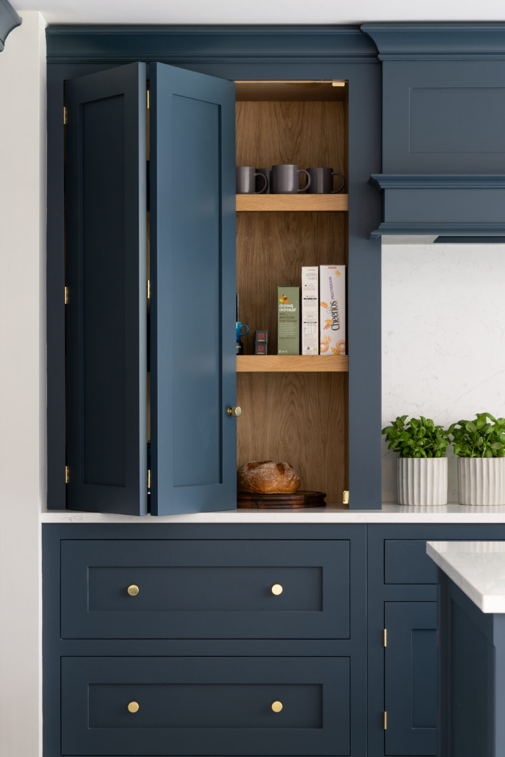 Open bi-fold cabinet doors reveal shelves with mugs, breakfast boxes and bread, set within deep blue cabinetry with brass handles