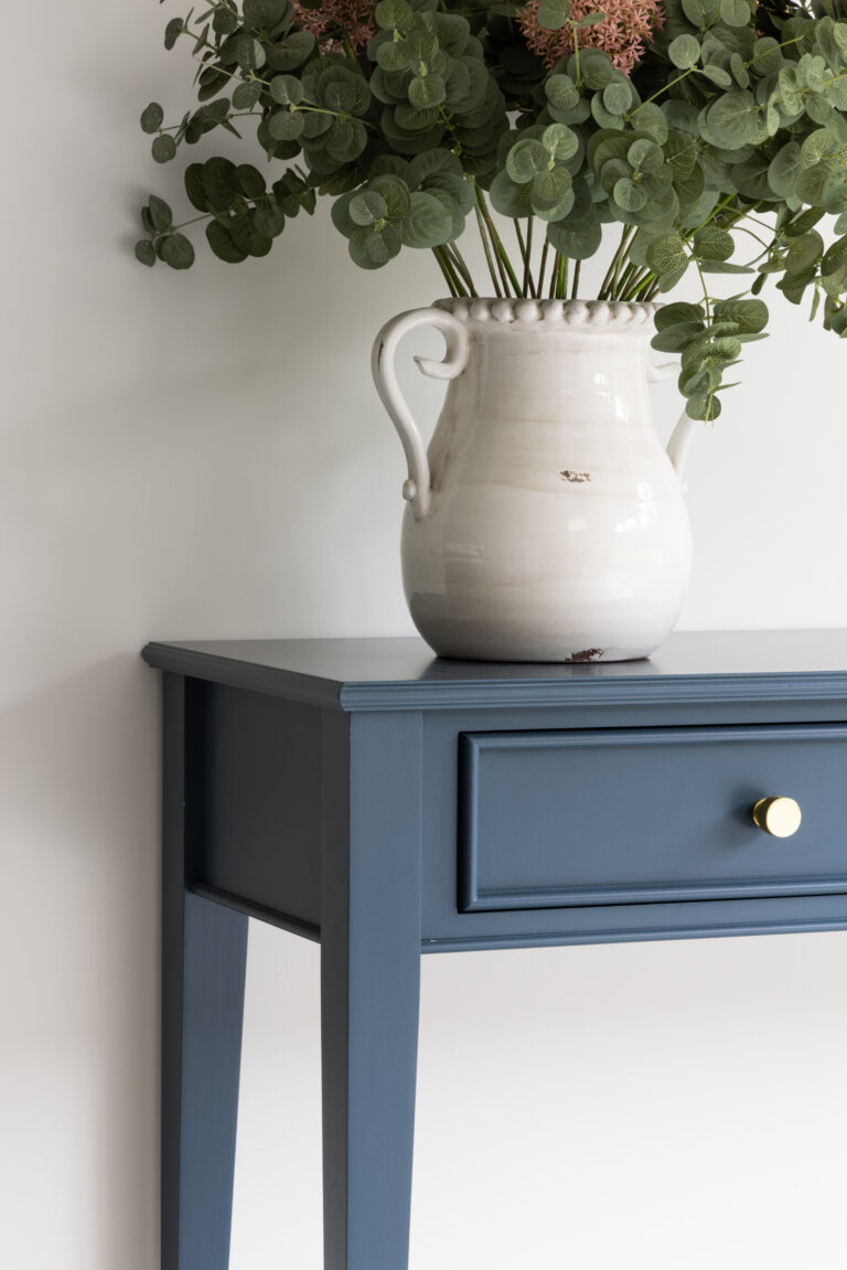 Blue side table with a brass drawer handle, topped with a ceramic jug holding eucalyptus foliage