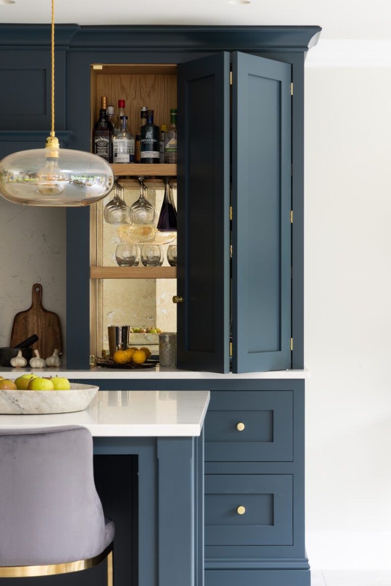 Open bi-fold Hague Blue cabinet revealing an oak-lined home bar with stemware racks, spirit bottles and mirrored splashback