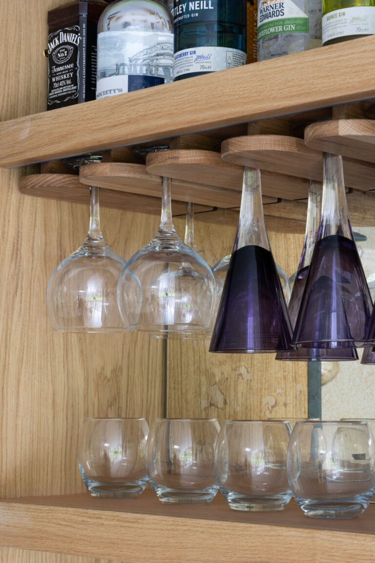 Close-up of an oak home bar interior with custom stemware racks holding glasses above spirit bottles and tumblers