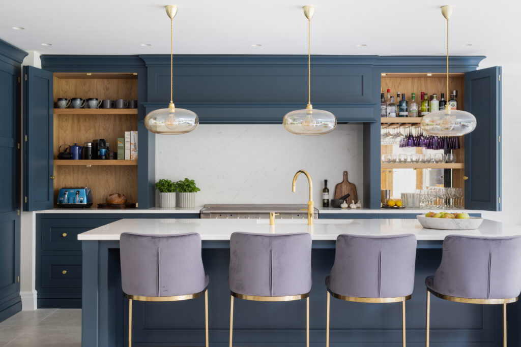 Open-plan kitchen with Hague Blue cabinetry, twin bi-fold units revealing an oak-lined bar and breakfast station, island with velvet stools
