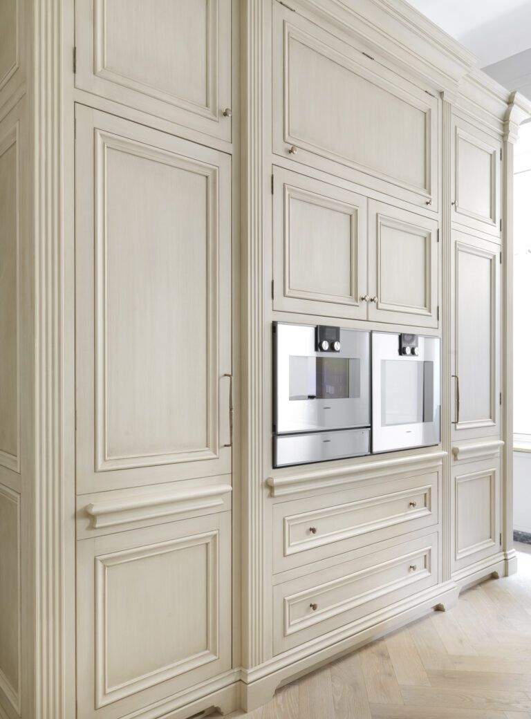 Tall hand-painted cabinetry with panelled doors and two integrated ovens above a herringbone timber floor