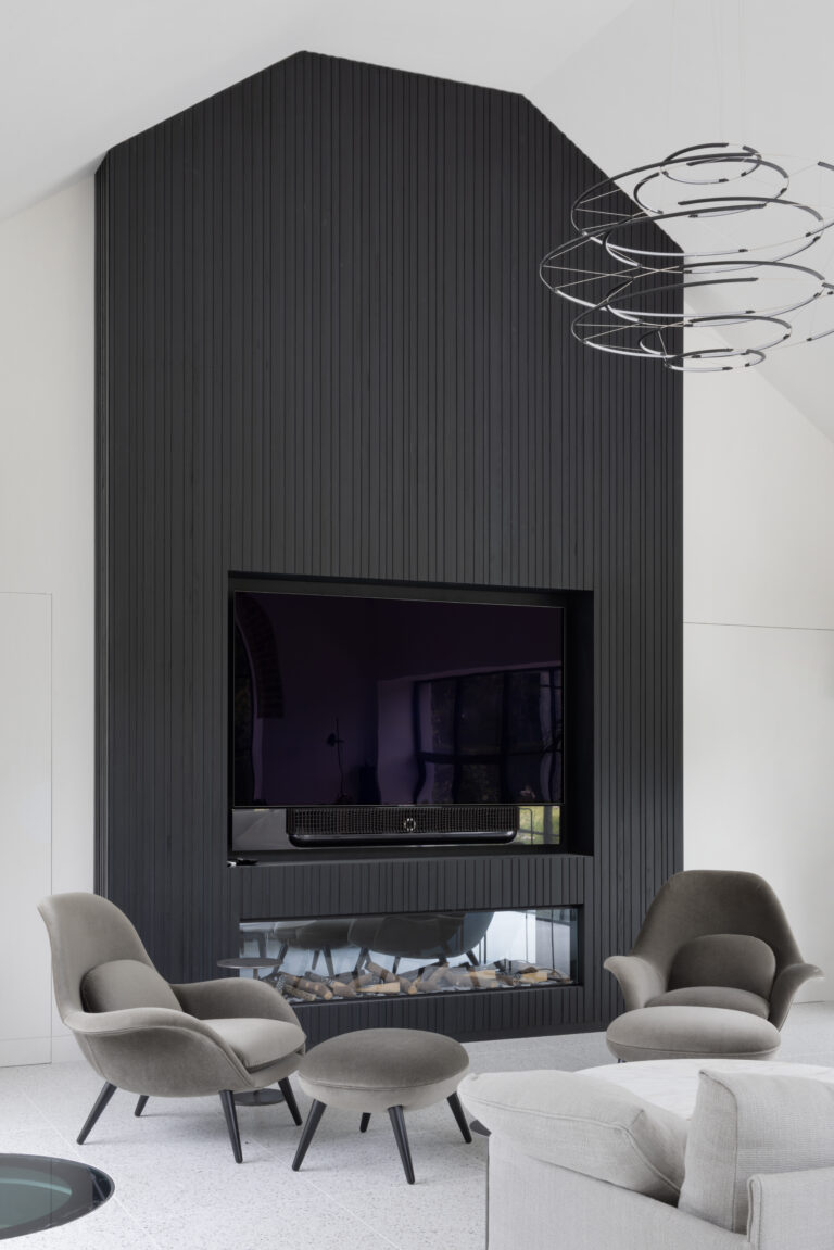 Double-height lounge with black slatted ash media wall housing a television above a built-in fireplace, flanked by low armchairs