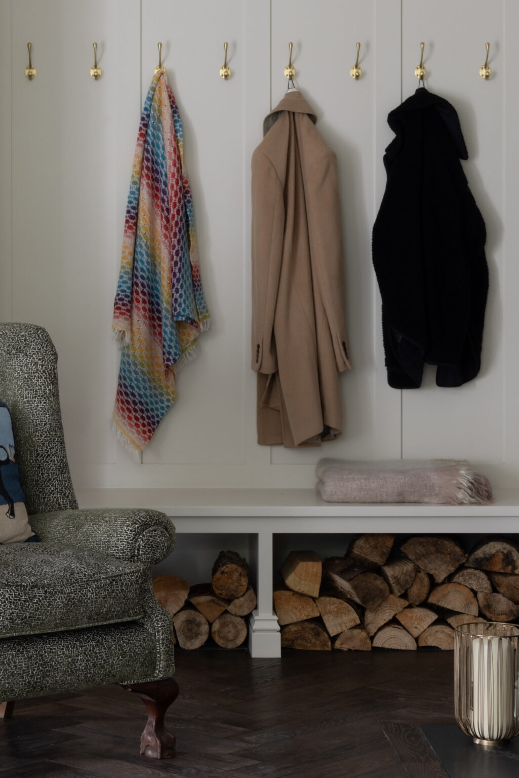 Boot room panelling with Satin Brass coat hooks, log storage beneath, and a patterned armchair beside the herringbone floor