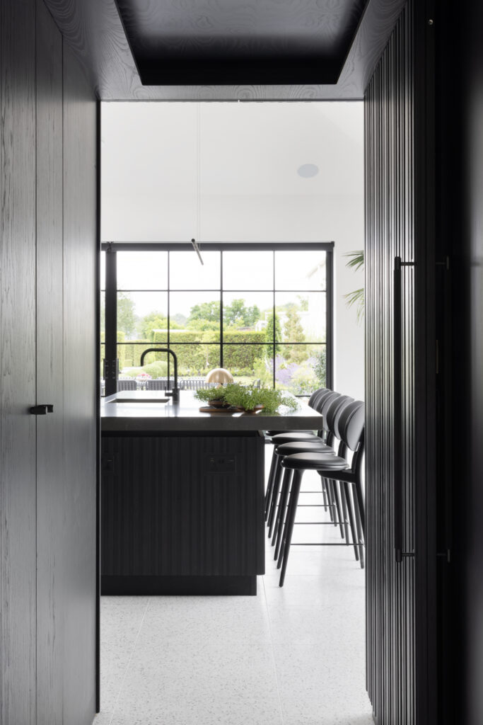 View through a doorway to a kitchen island with black bar stools, overlooking a crittall window onto the garden