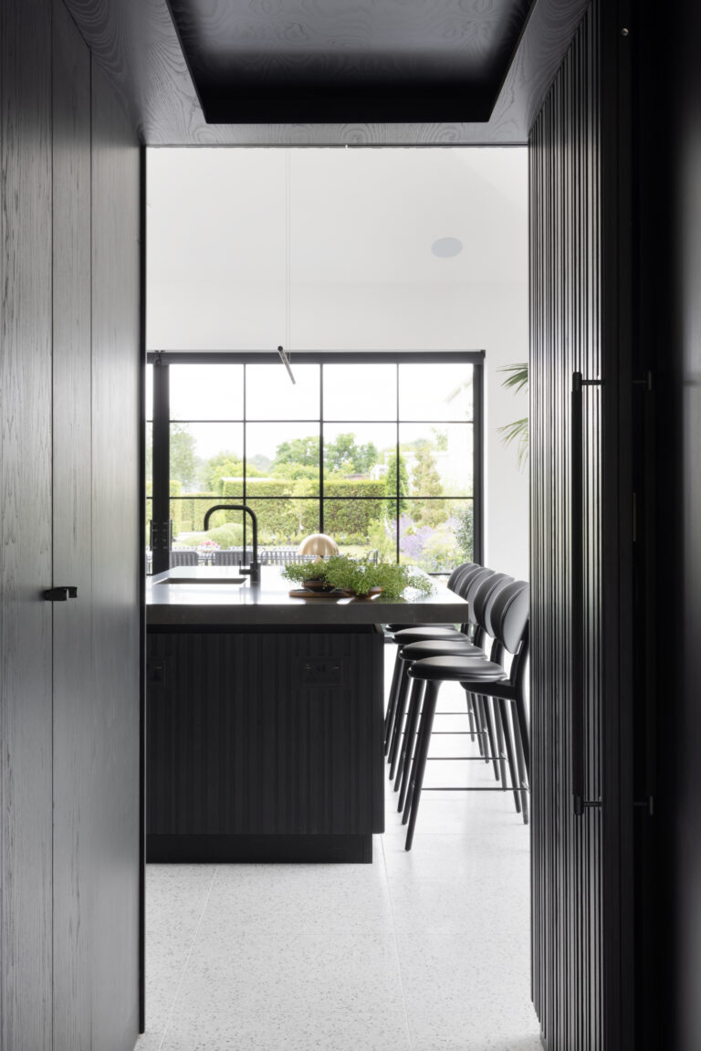 View through a doorway to a kitchen island with black bar stools, overlooking a crittall window onto the garden