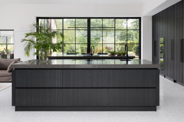 Large fluted dark kitchen island with stone worktop, black tap and sink, facing a crittall-style garden window