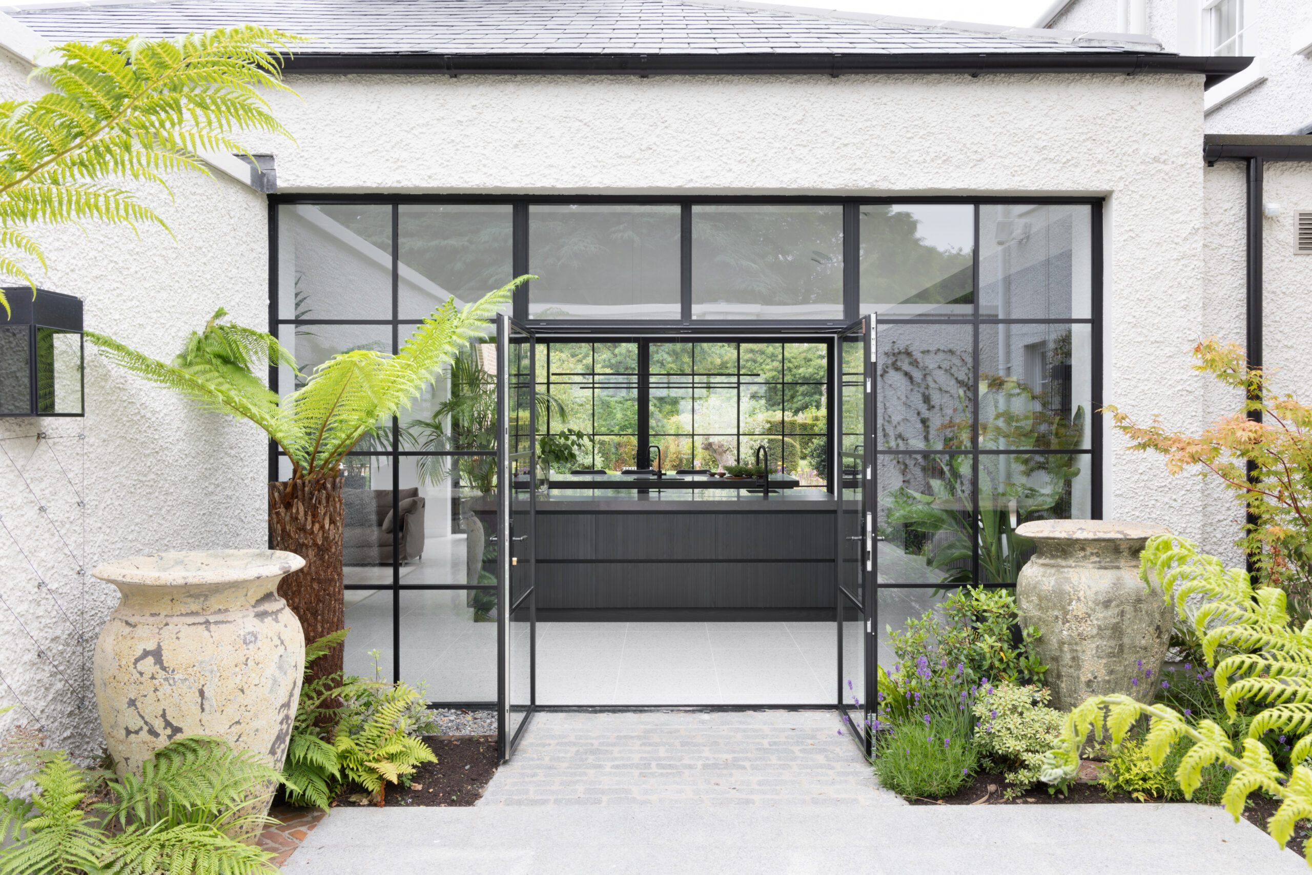 Crittall-style glazed doors open onto a dark kitchen island, framed by ferns and stone urns in the courtyard