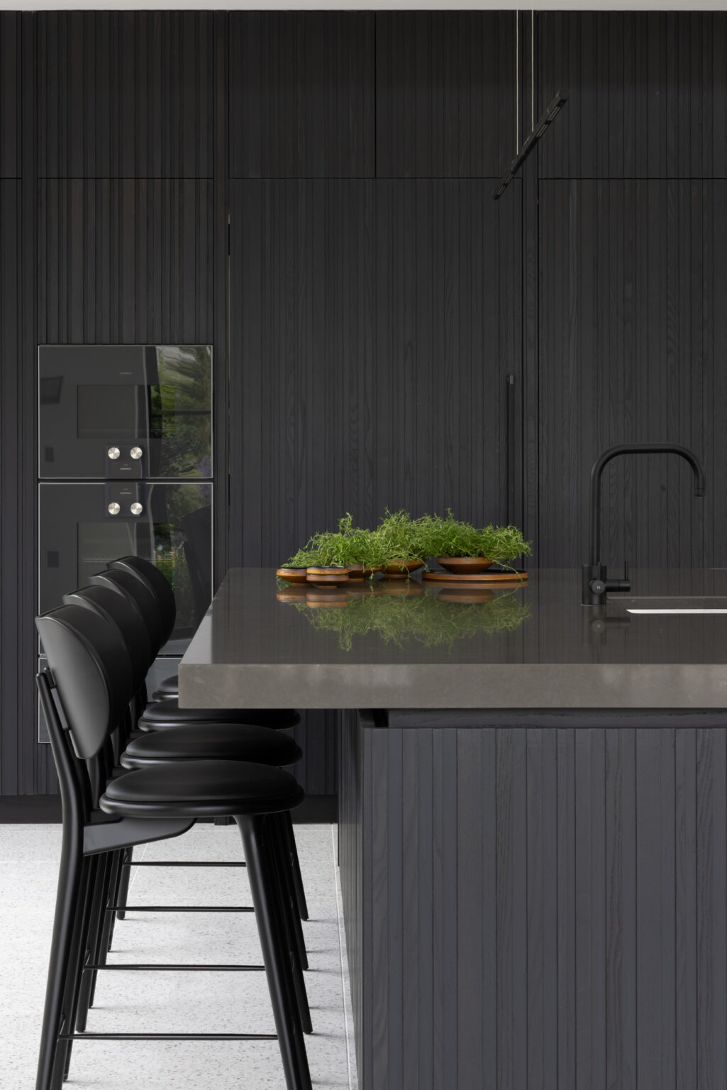 Dark fluted kitchen cabinetry with black bar stools, built-in ovens and a stone-topped island with black tap