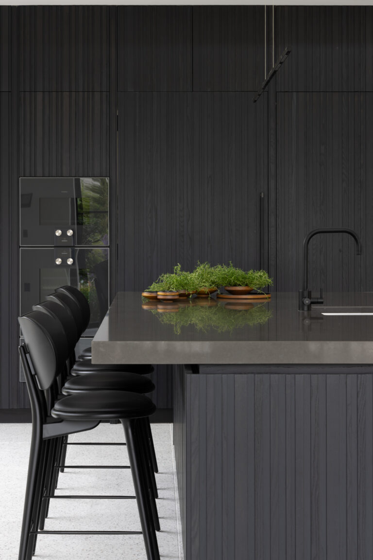 Dark fluted kitchen cabinetry with black bar stools, built-in ovens and a stone-topped island with black tap