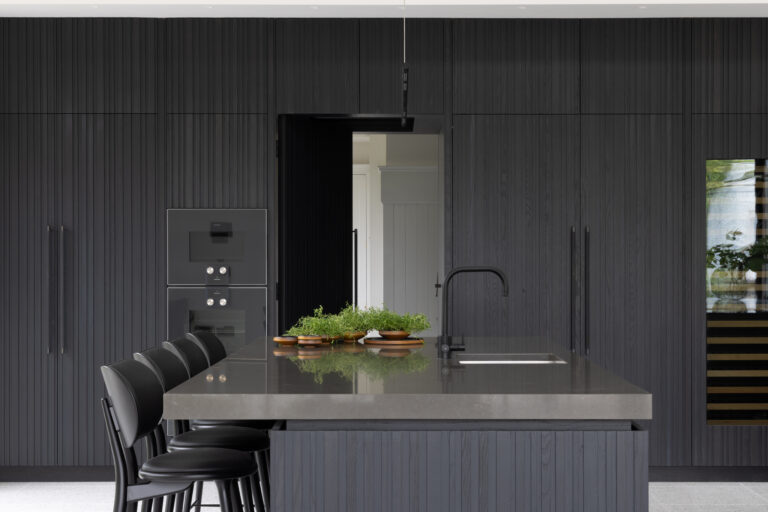 Dark fluted kitchen island with grey worktop, integrated sink, black tap, built-in ovens and bar stools