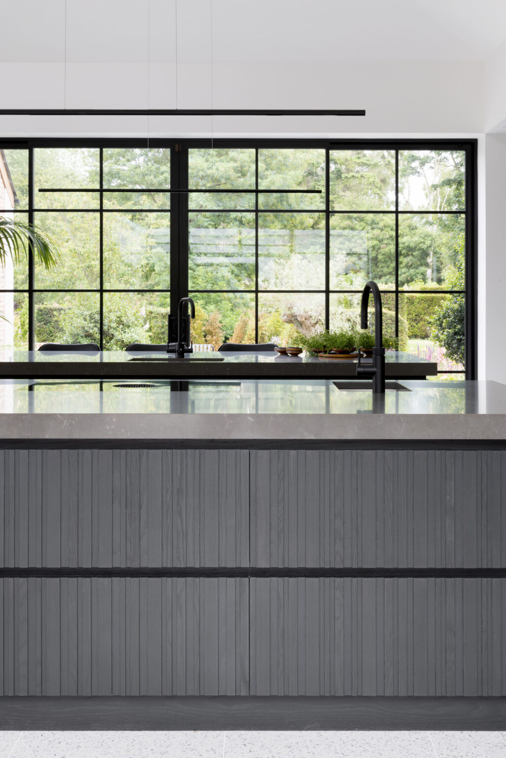 Ribbed kitchen island with black taps and stone worktop stands before a large Crittall-style window overlooking the garden
