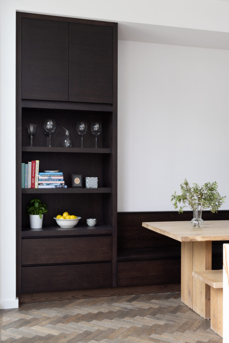 Dark built-in shelving unit displays glassware and books beside a pale wood dining table on herringbone flooring