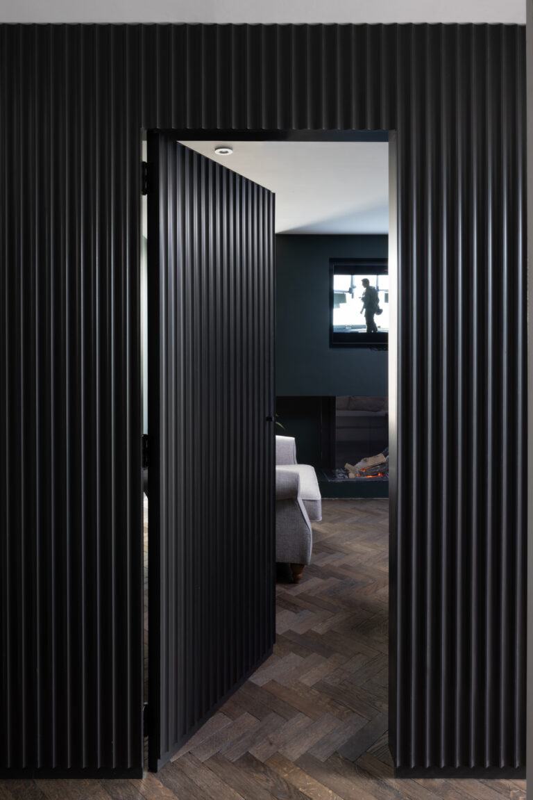 Fluted dark panelled door opens onto a snug with herringbone flooring, fireplace and sofa beyond