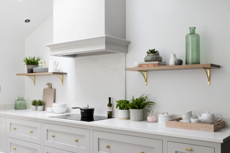 Pale grey cabinetry with brass handles beneath floating shelves, a cooker hood, induction hob and styled crockery