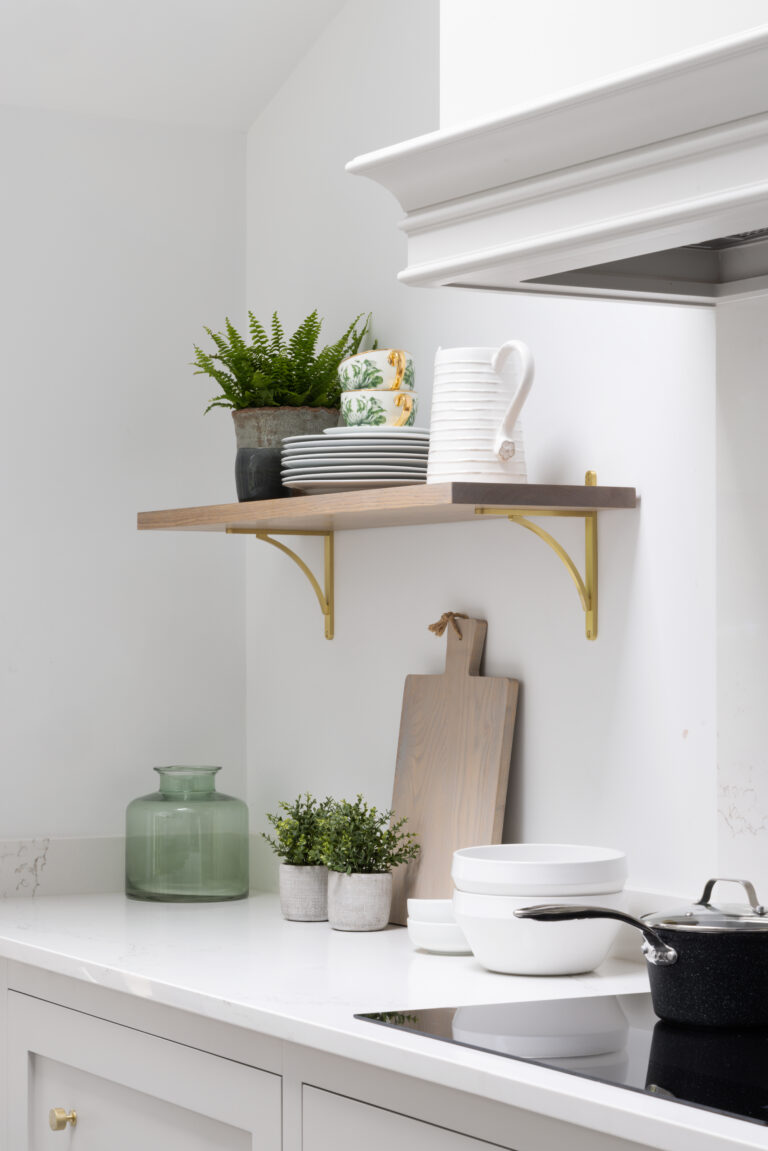 Floating shelf with brass brackets holds crockery and plants above a white worktop with a chopping board