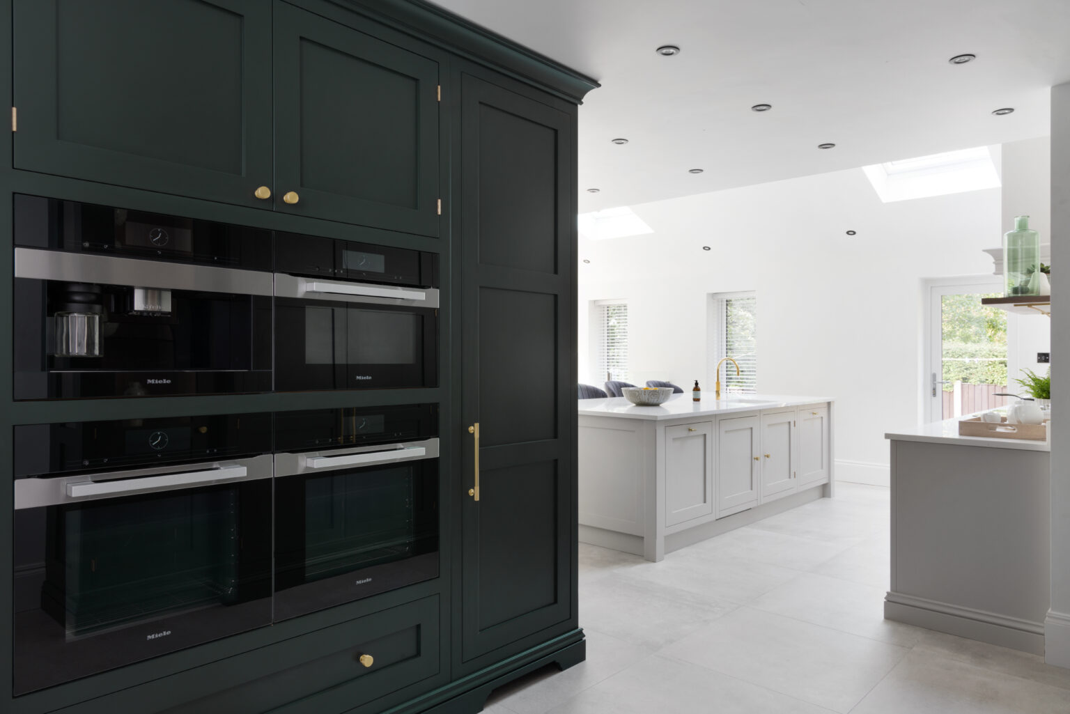 Dark cabinetry houses stacked Miele ovens with brass handles, framing a bright island beyond with skylights above