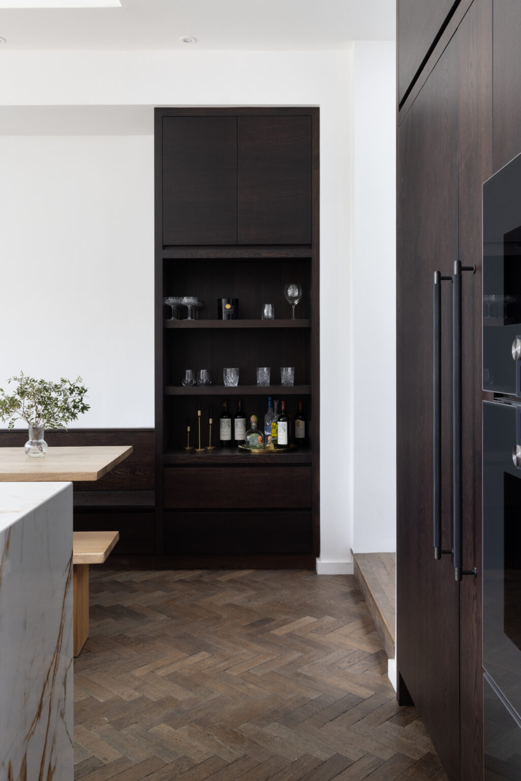 Dark built-in drinks cabinet with open shelving for glassware and bottles above herringbone parquet flooring
