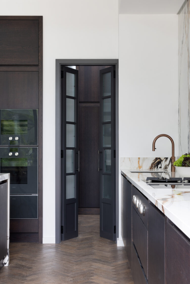 Glazed folding doors open to reveal the hidden walk-in pantry beside built-in ovens and a copper tap island