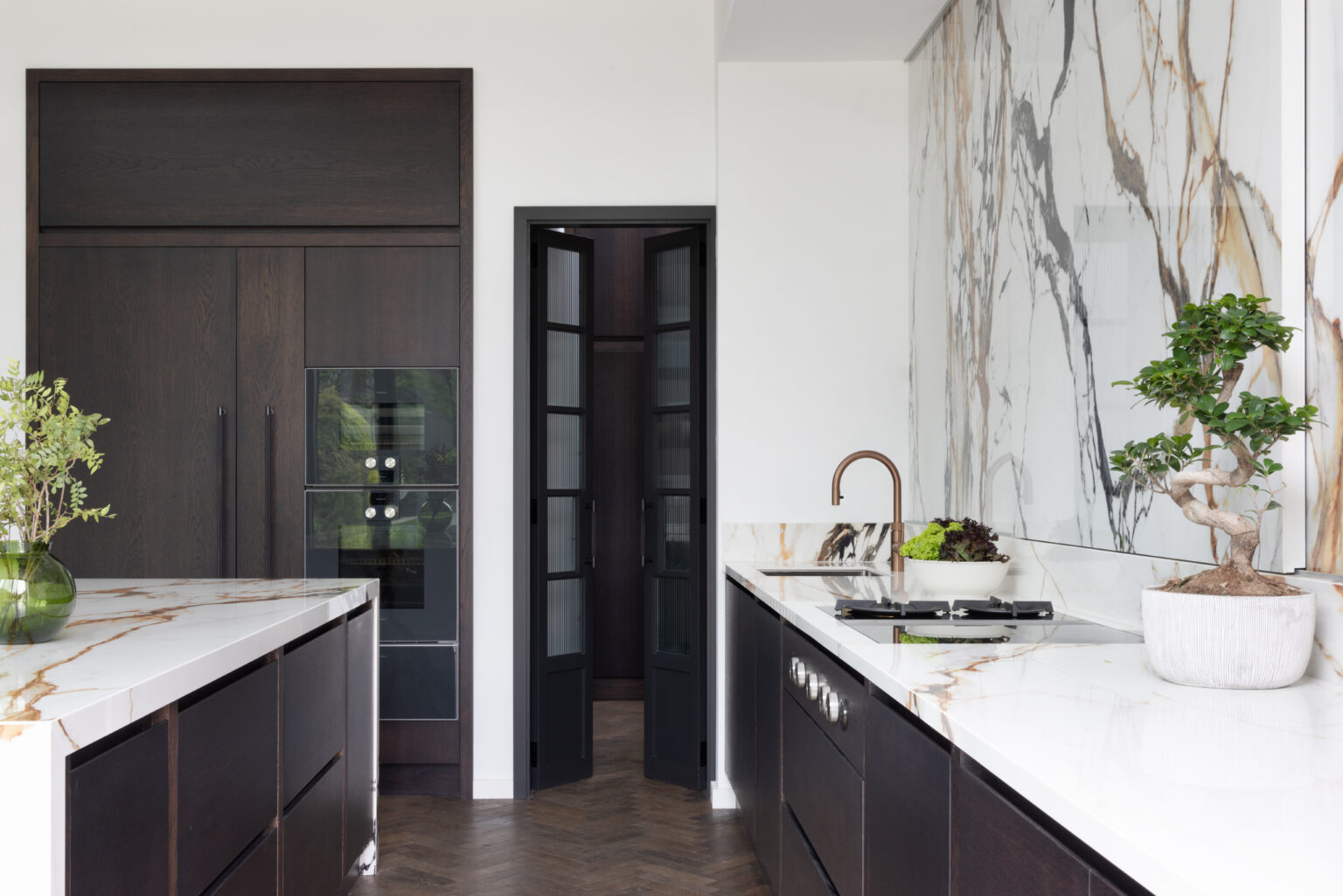 Open-plan kitchen with dark cabinetry, a moving stone splashback wall, copper tap and herringbone wood flooring