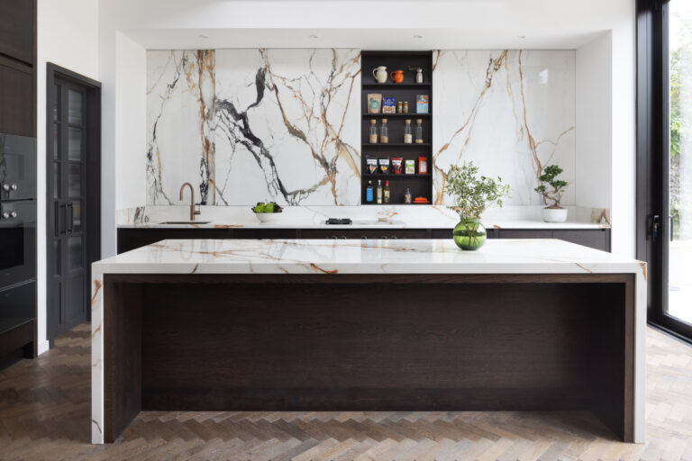 Open-plan kitchen island with dark timber panelling, veined stone worktop and matching wall of moving stone behind