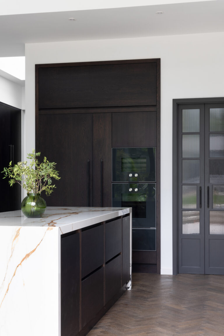 Open-plan kitchen with dark cabinetry, integrated ovens, veined stone island top and reeded glass pantry doors