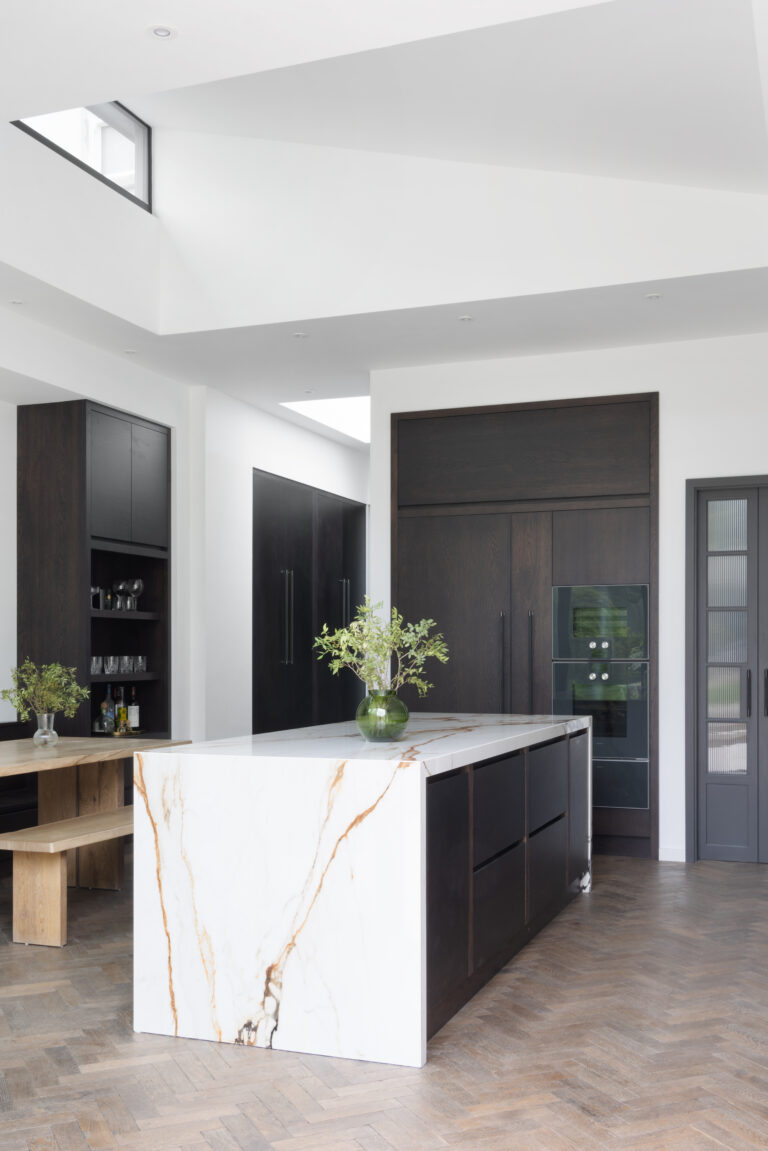 Open-plan kitchen with a veined stone island, dark cabinetry, built-in ovens and herringbone wood flooring