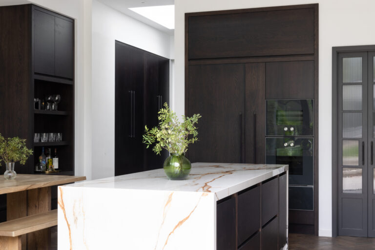 Kitchen island with a veined waterfall stone worktop, dark cabinetry, built-in wine fridge and glassware nook behind
