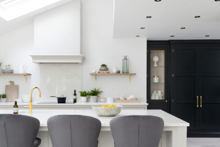 Kitchen island with grey bar stools, white countertops, open shelving and dark cabinetry with brass handles beyond