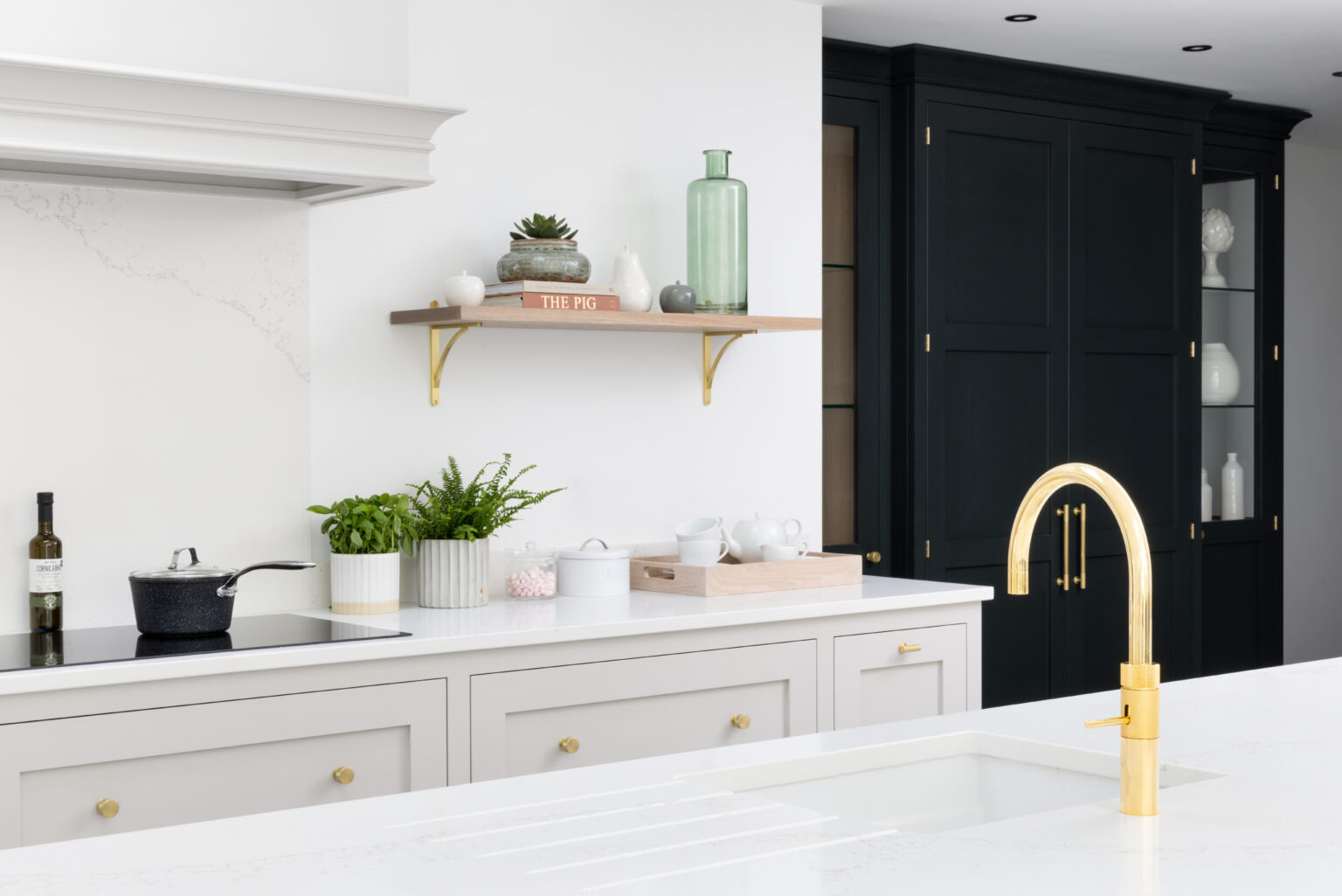 Light cabinetry with brass handles sits beside dark cabinetry, a floating shelf and gold tap defining the colour-blocked kitchen