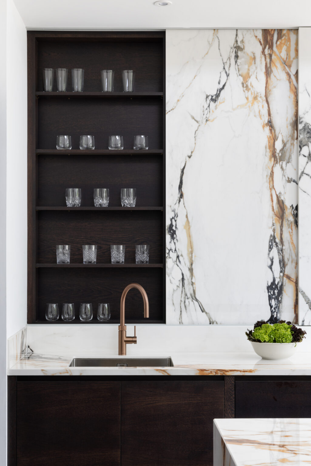 Open shelving displays glassware beside a sink with copper tap, set against a dramatic veined stone wall panel