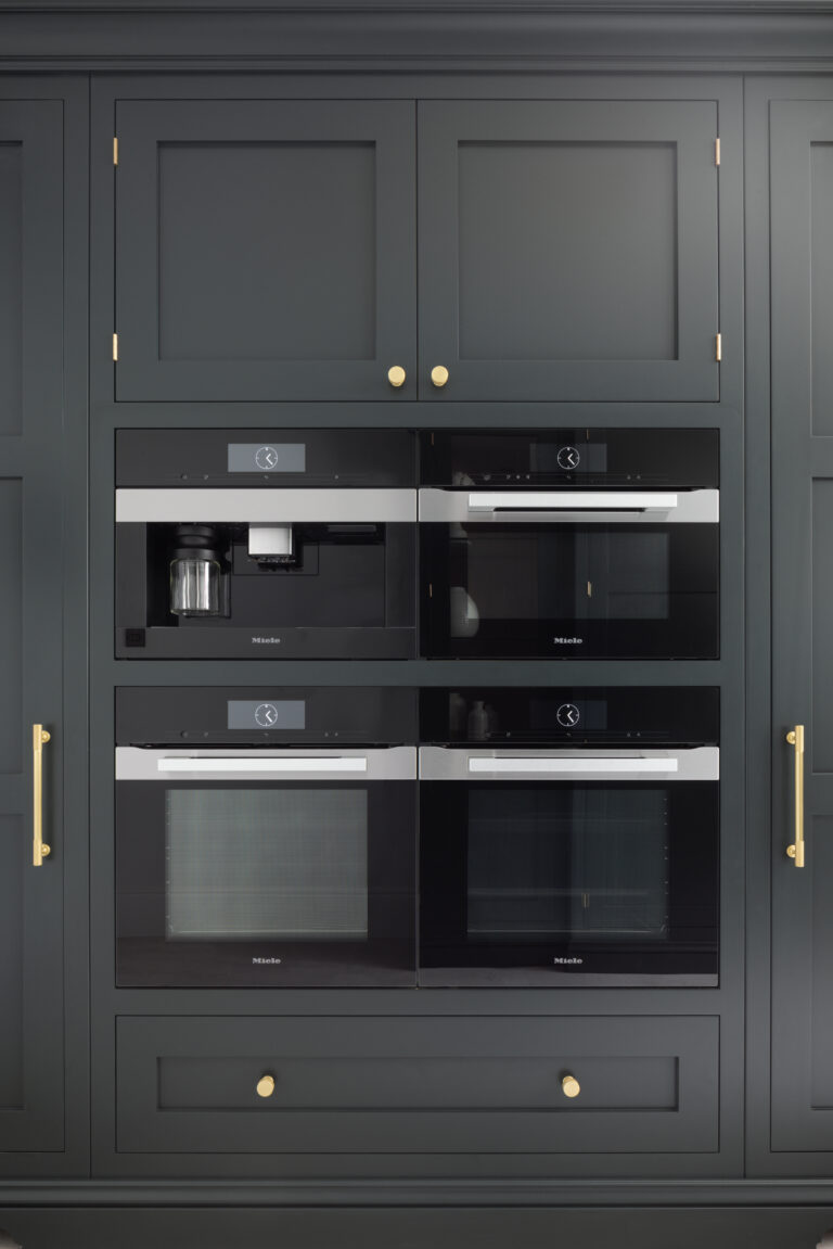 Dark cabinetry houses four stacked Miele appliances, including a coffee machine, finished with brass handles and knobs