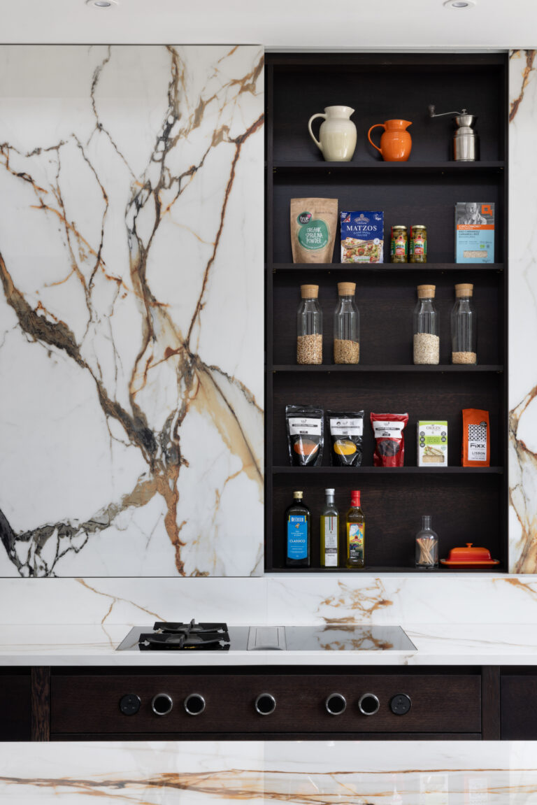 Open shelving beside a gas hob displays jars, oils and pantry items against a dramatic veined stone wall