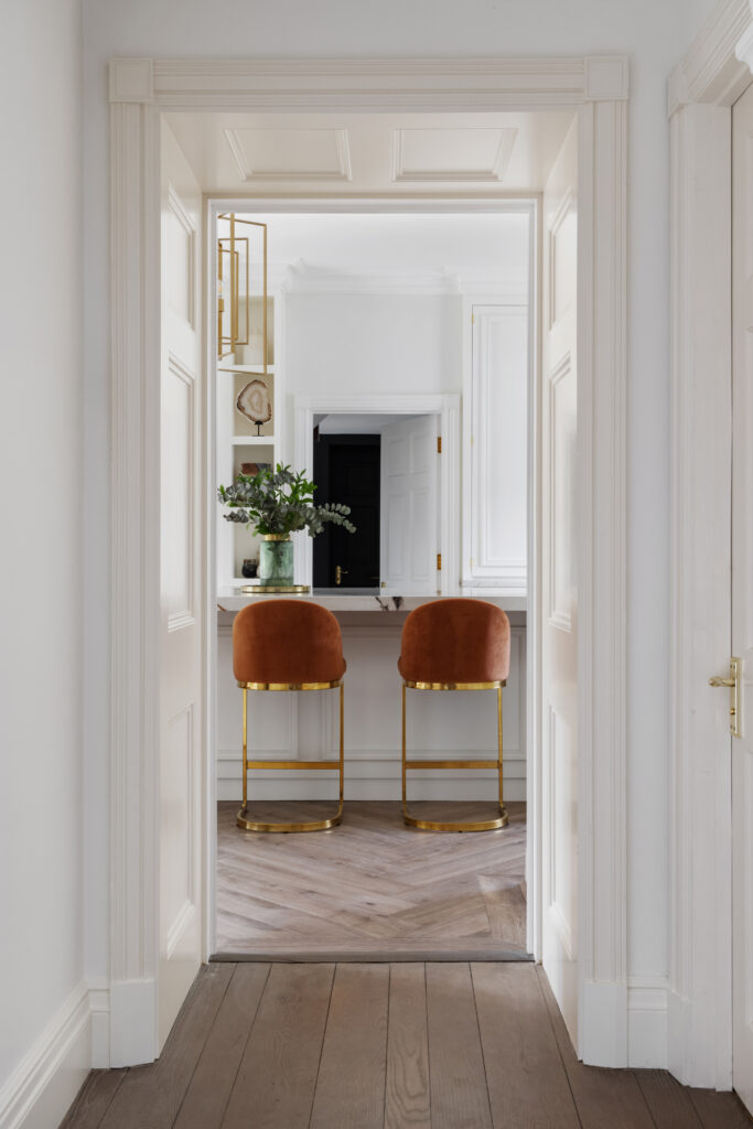 Hallway view through panelled white doors to burnt-orange velvet stools at a brass-legged kitchen island