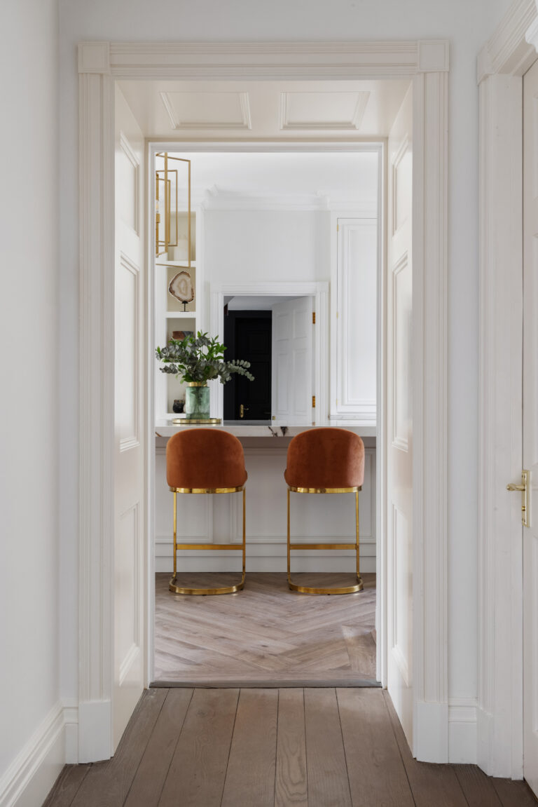 Hallway view through panelled white doors to burnt-orange velvet stools at a brass-legged kitchen island