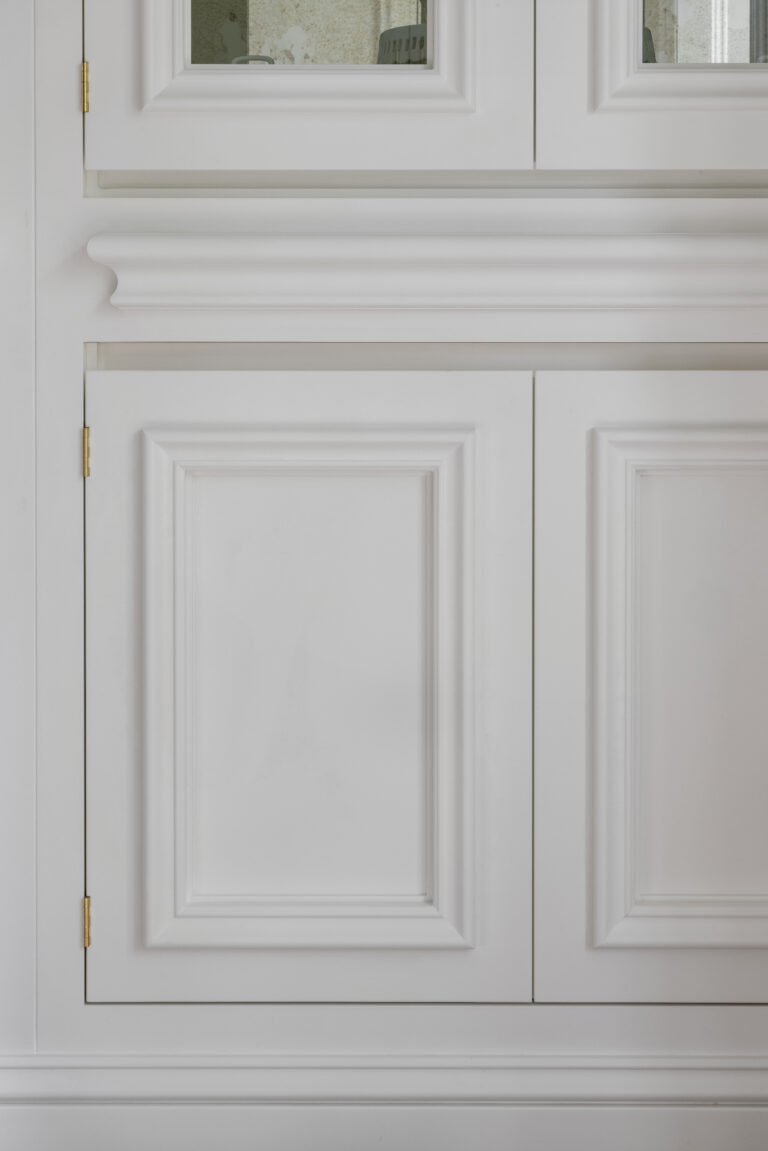 Close-up of white panelled cabinetry with brass hinges, moulded cornicing and small antique mirrored door panels