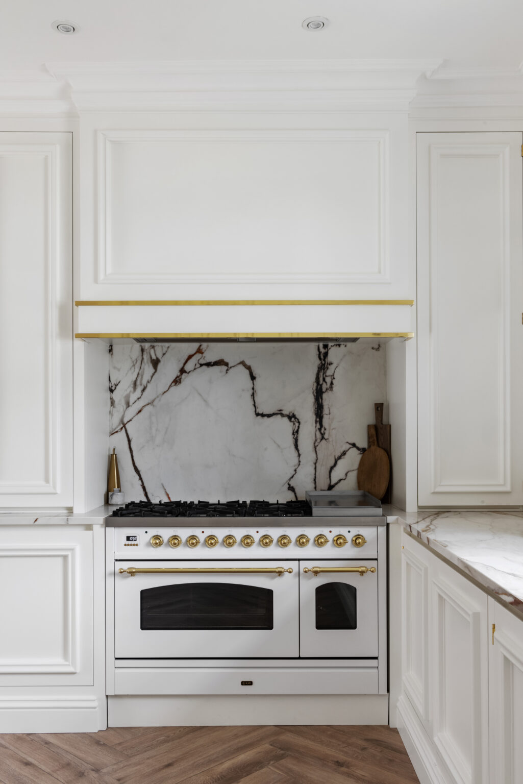 White range cooker with brass fittings set within panelled white cabinetry and a veined stone splashback