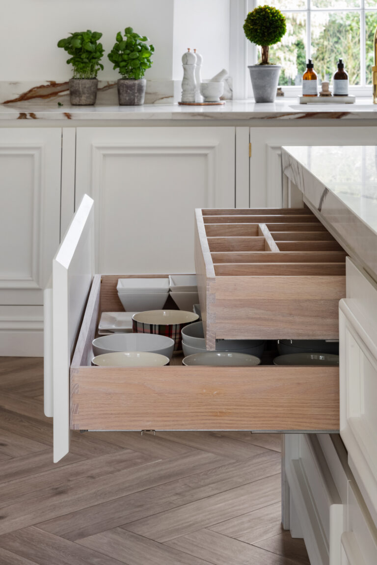 Open kitchen drawer with internal wooden organiser and stacked bowls, beneath a stone-topped island with herb pots