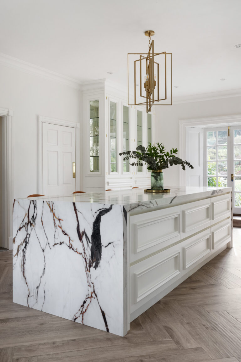 White kitchen island with a golden-veined XTONE waterfall edge, beneath a brass pendant light and glazed display cabinets