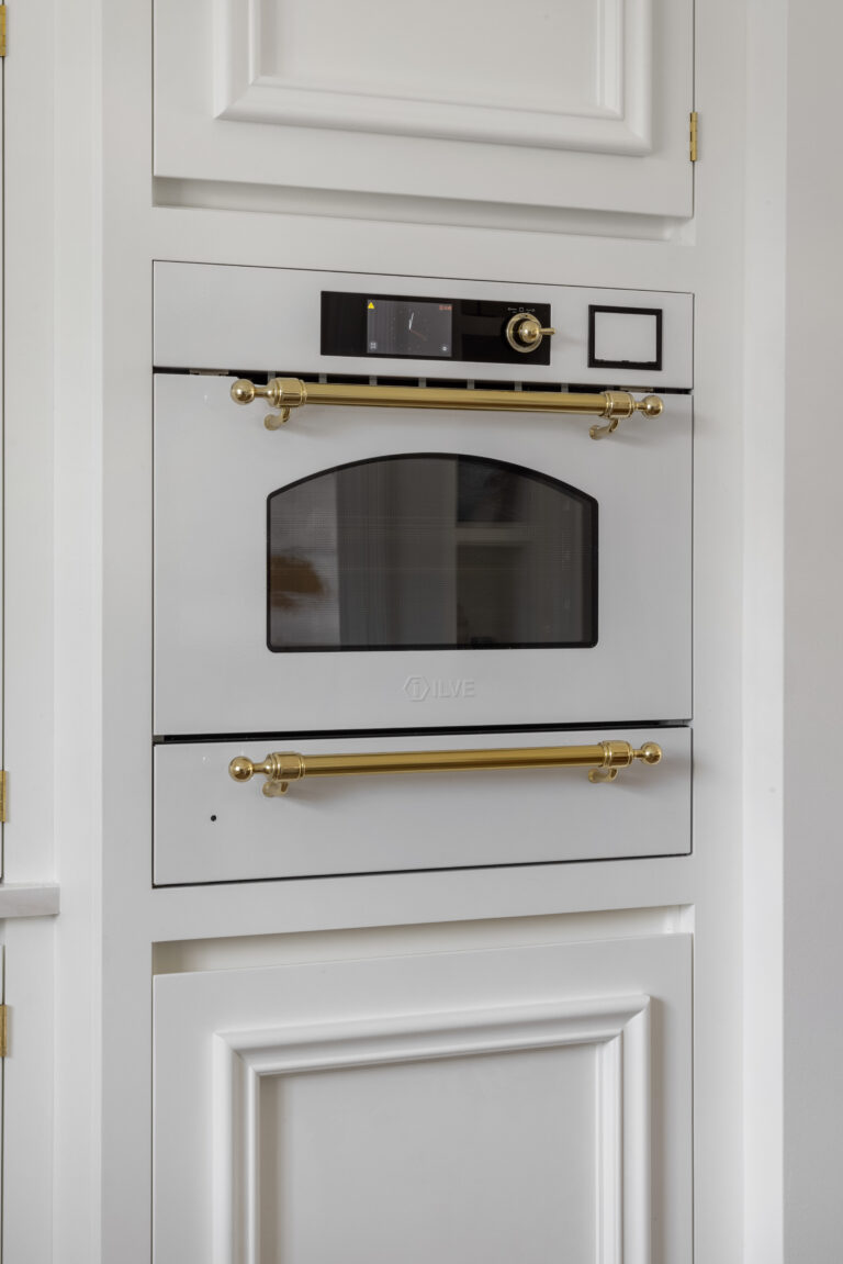 Built-in white oven with brass handles and arched glass door, set within panelled white cabinetry