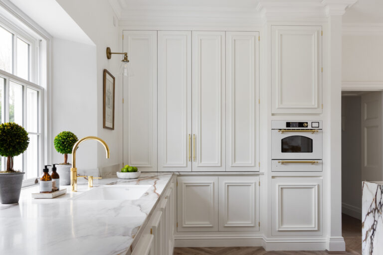 White panelled cabinetry with brass handles, a gold tap and veined XTONE island worktop by the window