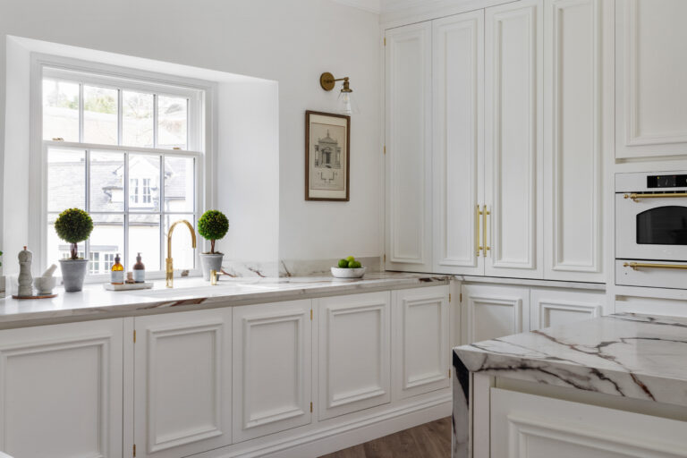White panelled cabinetry with brass handles surrounds a window, with an XTONE island featuring golden veining nearby