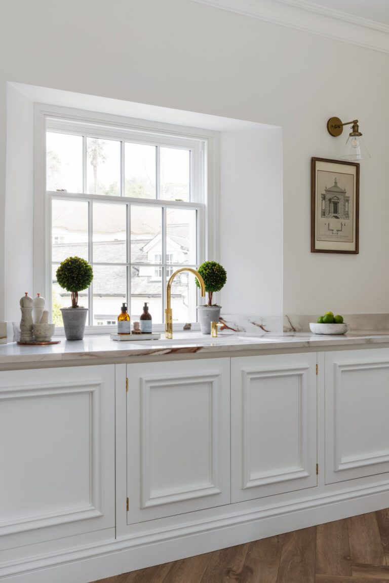White panelled cabinetry beneath a sash window, with a brass tap, veined worktop and brass wall light